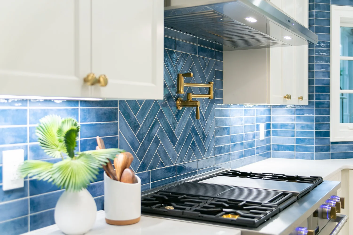 Ford’s Colony kitchen cooking wall remodeled by VanFleet Homes with white cabinetry, a blue herringbone backsplash, brass pot filler, and professional range