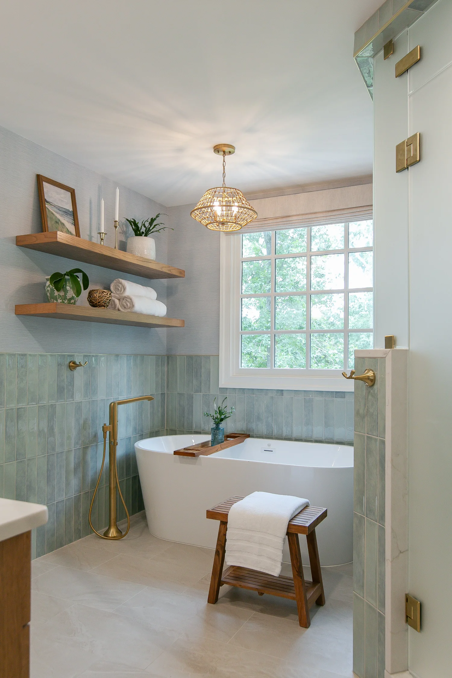 Completed Ford’s Colony bathroom remodeled by VanFleet Homes with a freestanding tub, blue-green tile wainscoting, natural wood shelves, a woven pendant, and warm brass fixtures