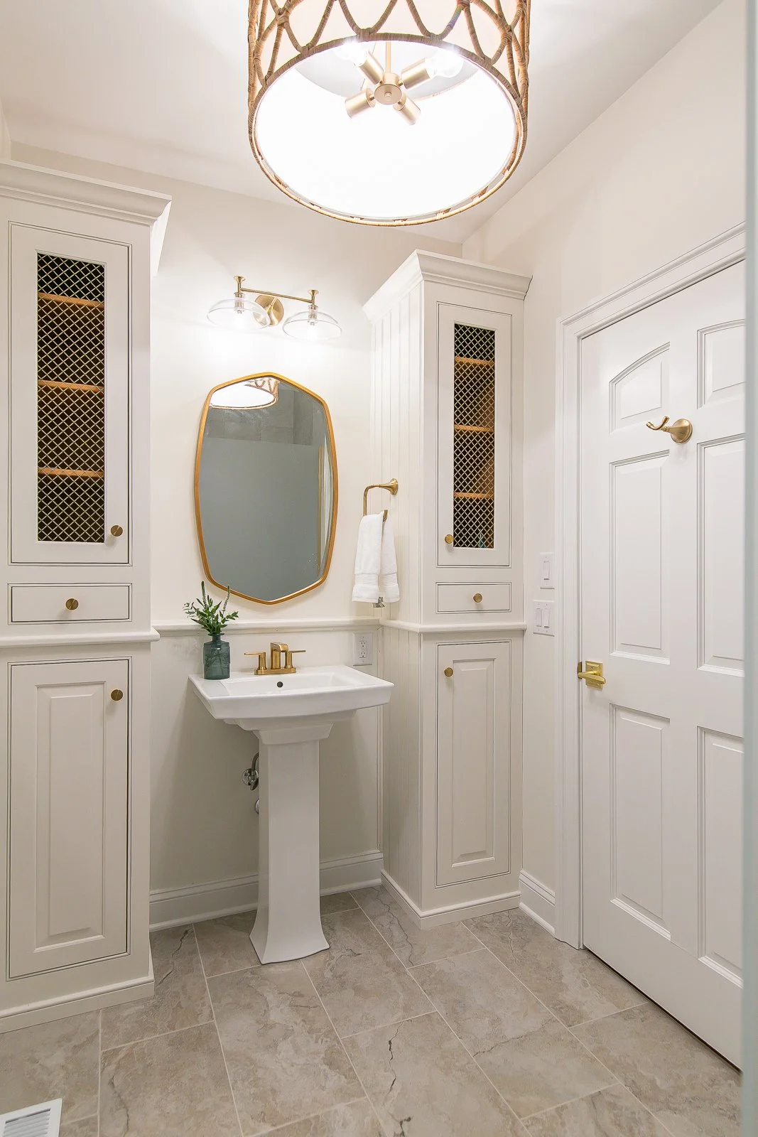 Completed Ford’s Colony powder room remodeled by VanFleet Homes with white built-in cabinetry, a pedestal sink, warm brass fixtures, a gold-framed mirror, and a woven drum light