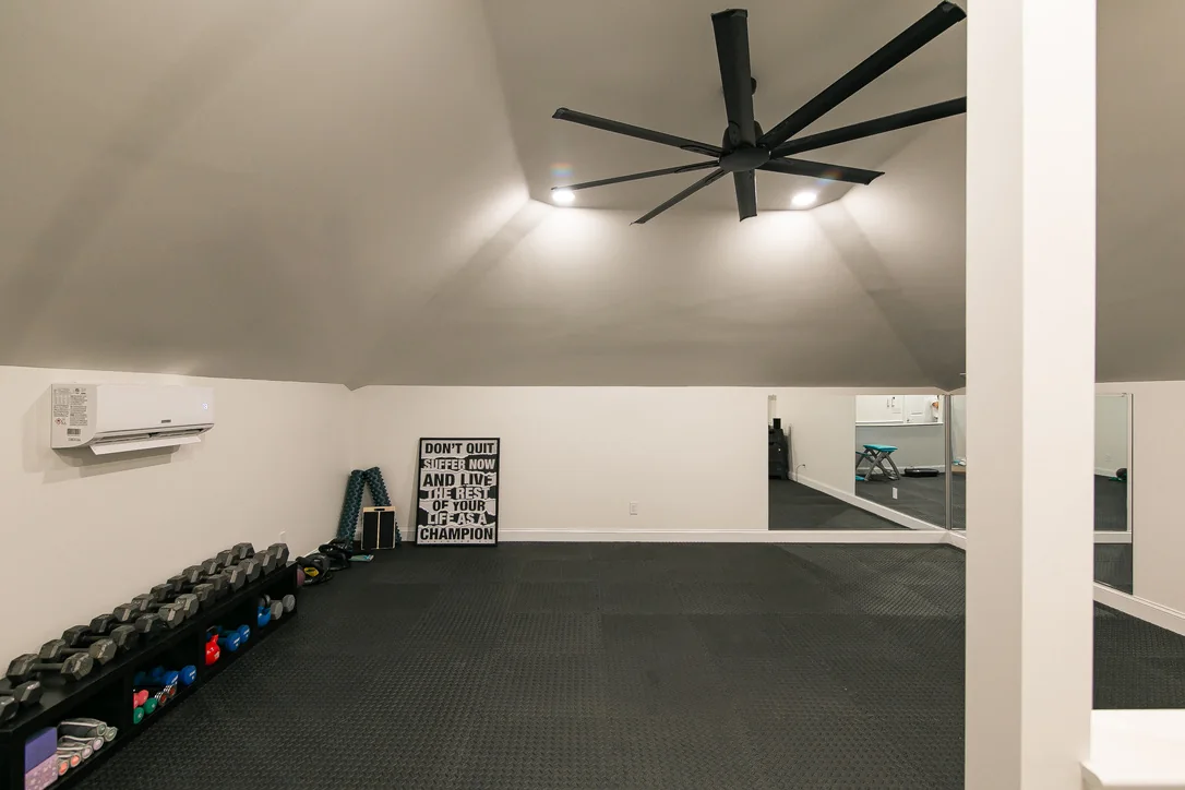 Completed attic home gym with gym flooring, wall mirrors, a large black fan, and mini-split