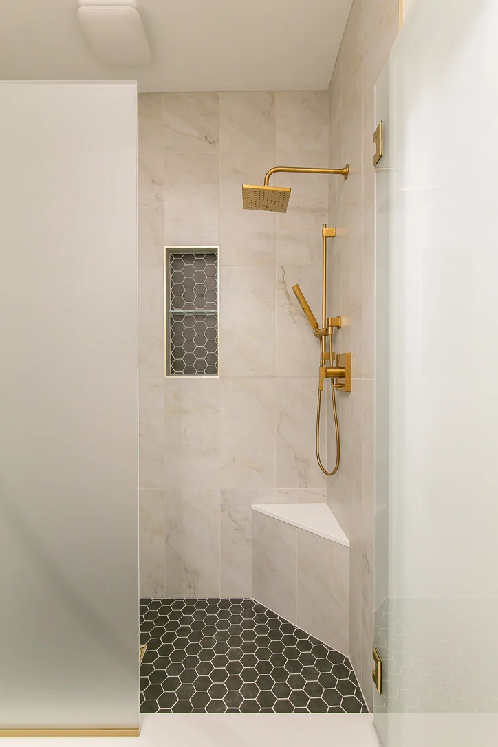 Completed guest walk-in shower with frameless glass, gold-toned fixtures, corner bench, niche, and dark hexagonal floor tile