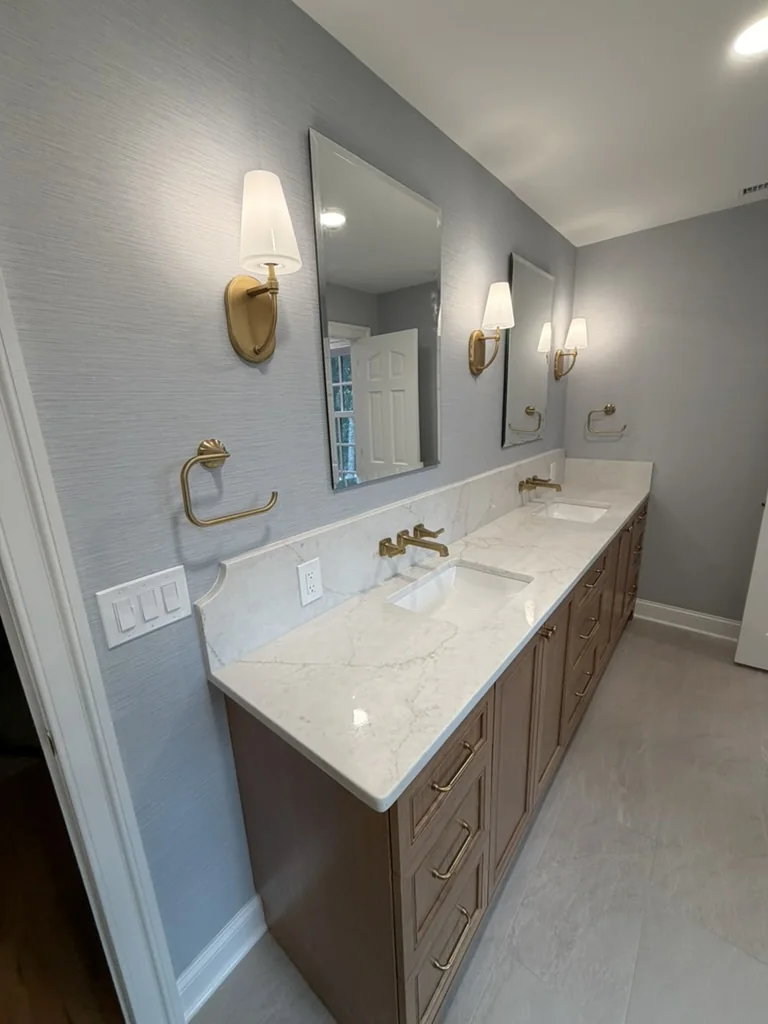 Completed stained white oak vanity with quartz backsplash, wall-mounted brass faucets, wallpaper, and sconces