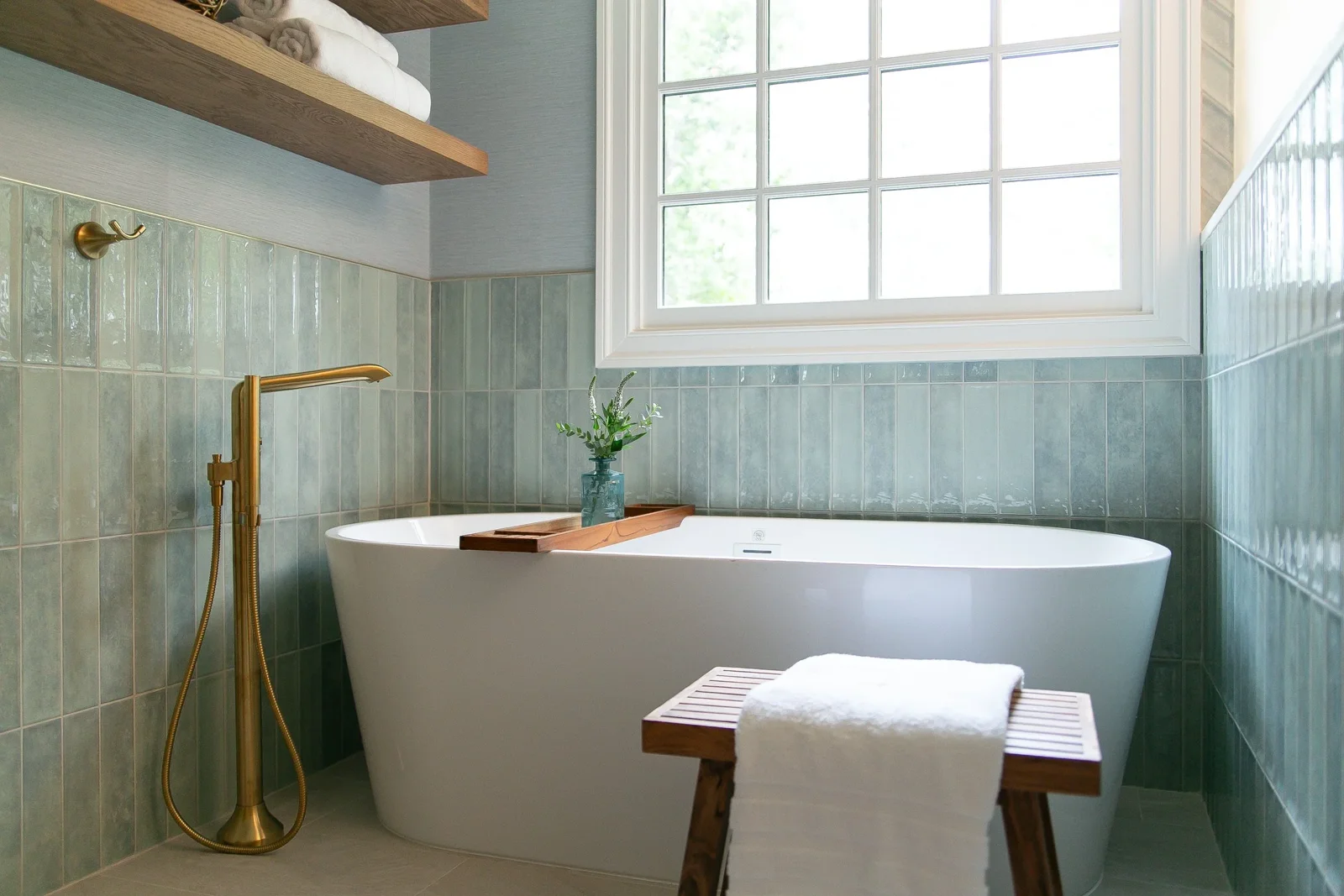 Finished freestanding tub beneath a window with blue-green tile and floating wood shelves