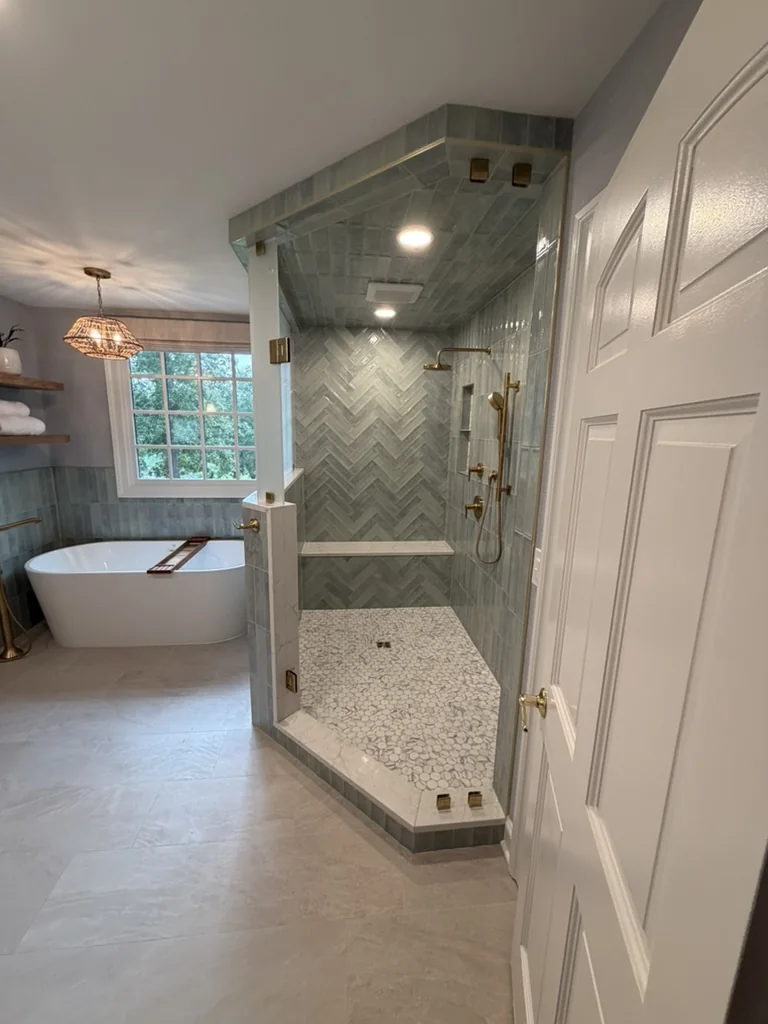 Completed custom shower with herringbone tile, pebble floor, bench, brass fixtures, and frameless glass