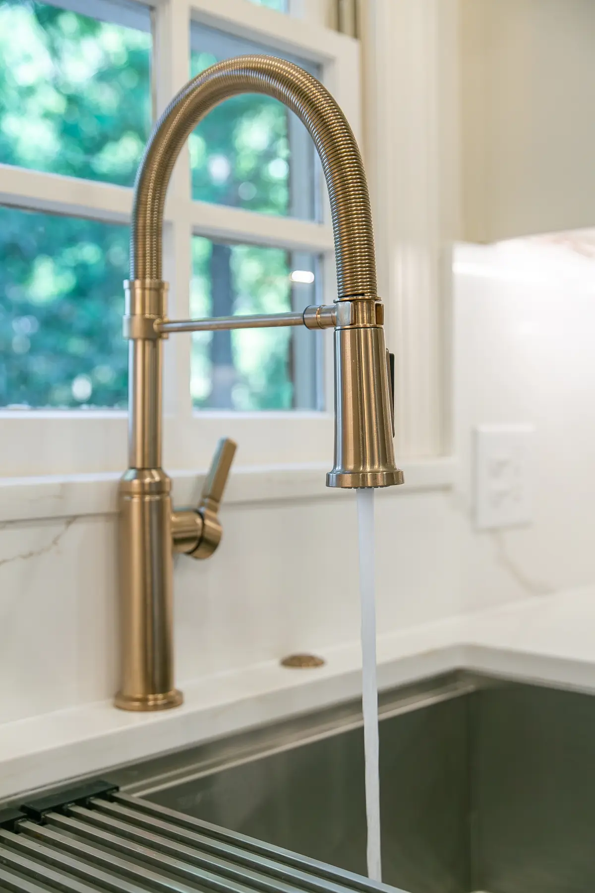 Brushed brass workstation faucet and stainless sink detail beneath a divided-light window