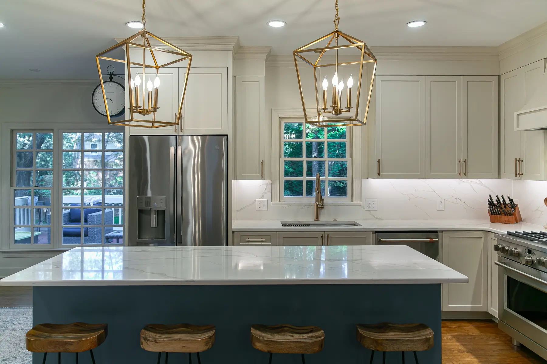 Front view of the blue kitchen island with four wood stools, brass lantern pendants, professional appliances, and warm white cabinetry in Kingsmill