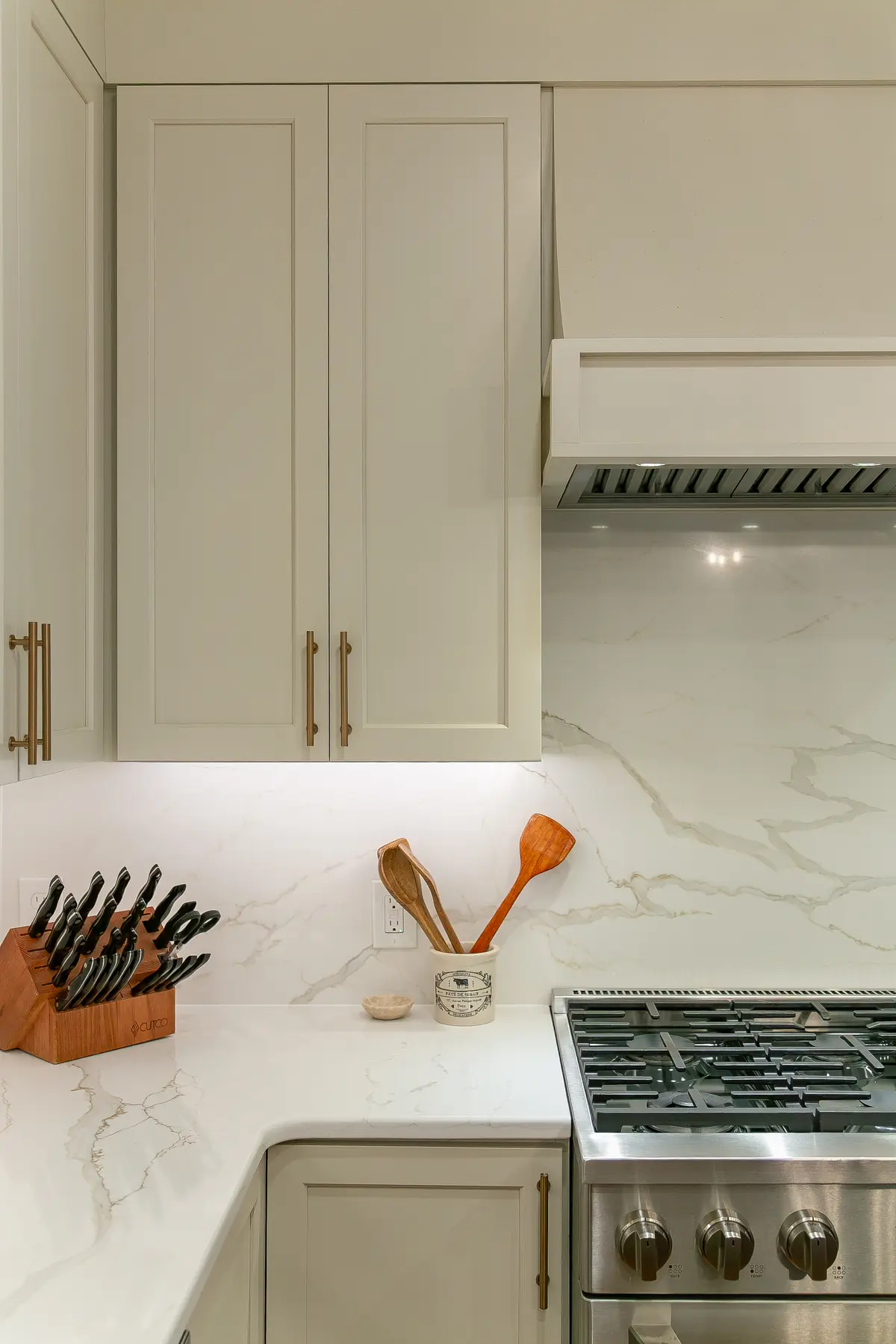 VanFleet Homes Kingsmill kitchen with a softly veined full-height slab backsplash, warm white shaker cabinets, brass pulls, and a stainless range beneath a tailored hood