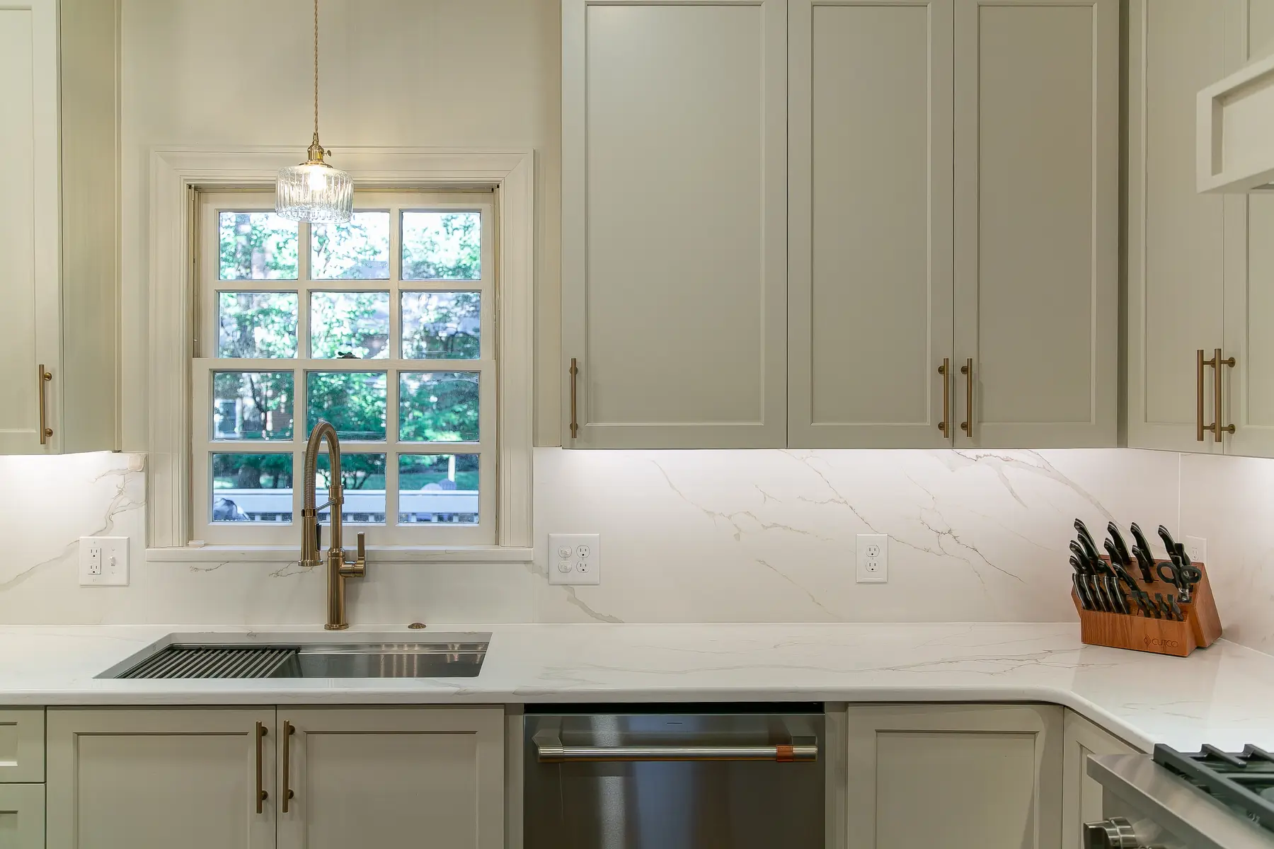 Symmetrical sink wall with warm white cabinetry, brass faucet, stone backsplash, dishwasher, and wooded Kingsmill view